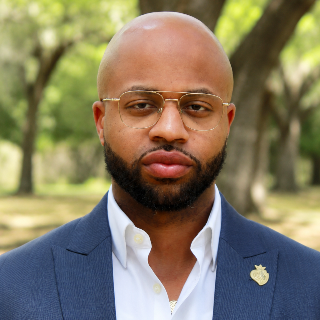 Bearded man wears a blue suit and white collared shirt with a lion head lapel pin